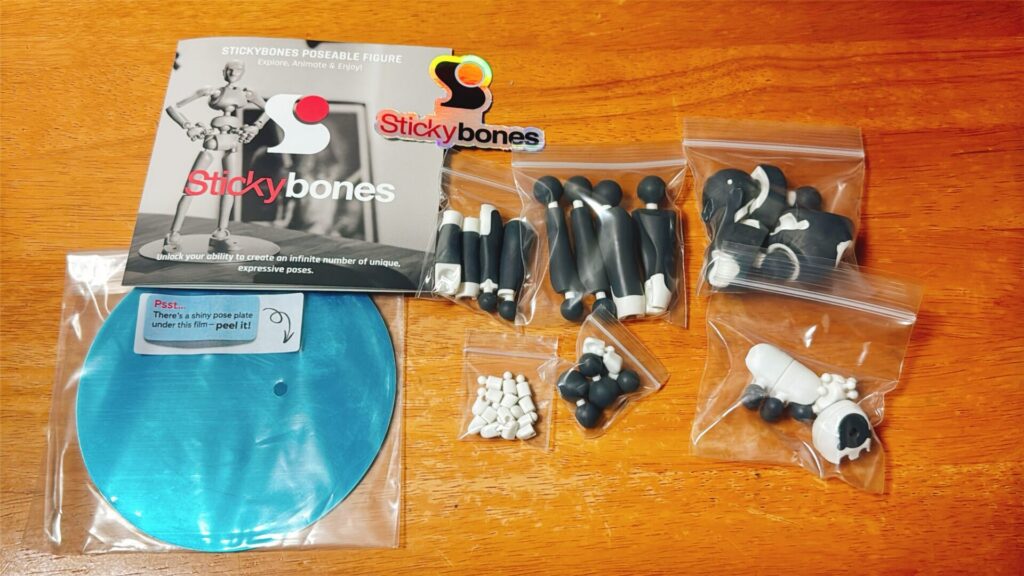 Stickybonesのパーツ一式が木のテーブルに並べられている。袋に入った黒と白の部品、青いポーズプレート、説明カードが写っている。