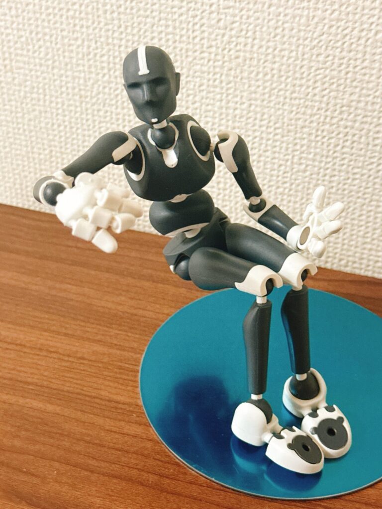 Stickybonesのフィギュアが青い台座の上で腰を下ろし、片腕を前に伸ばしたポーズをしている。木のテーブルと淡い壁を背景にした動きの確認用の姿勢。